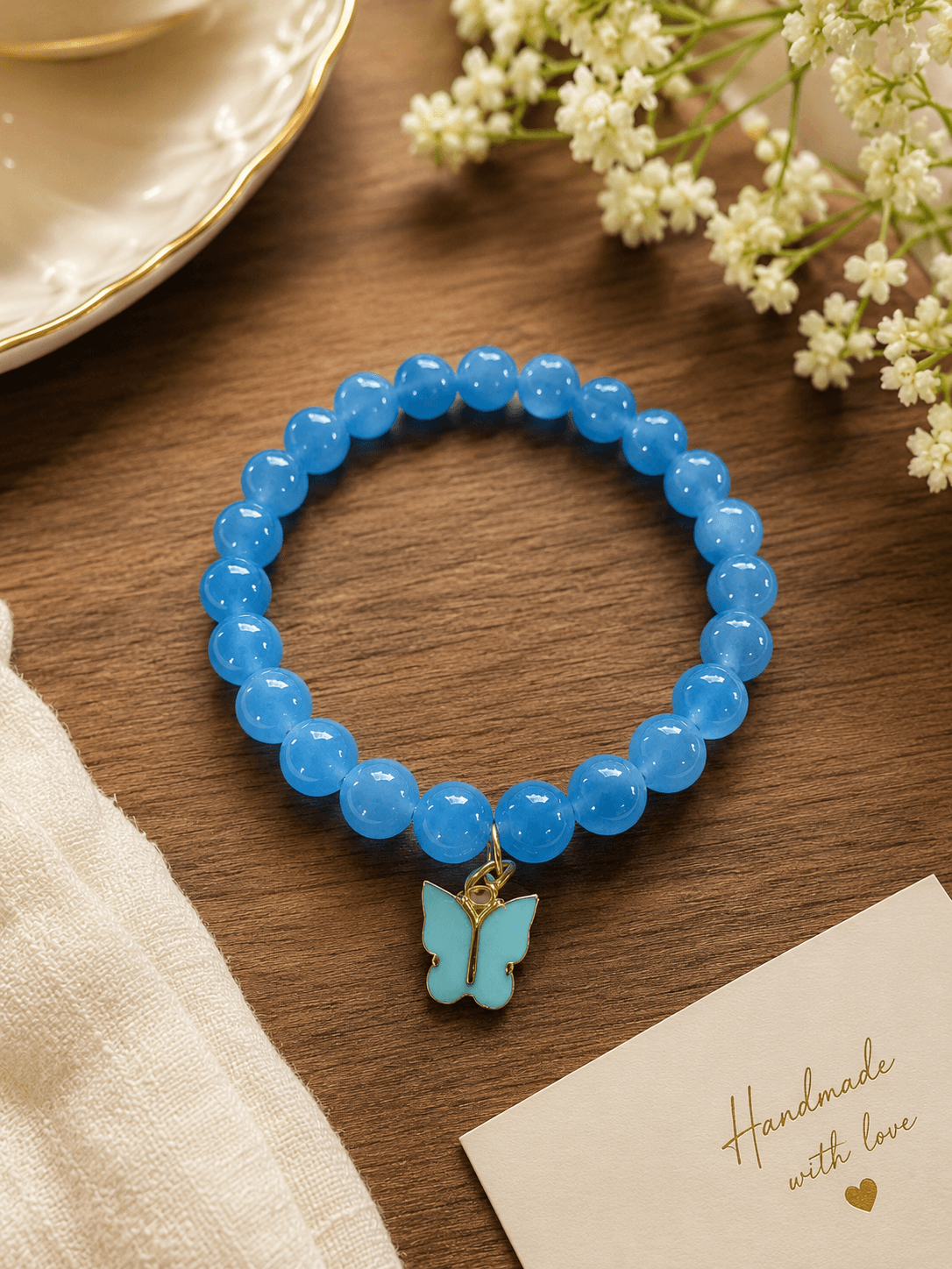 Handmade blue jelly bead stretch bracelet with a butterfly charm resting on wood next to flowers