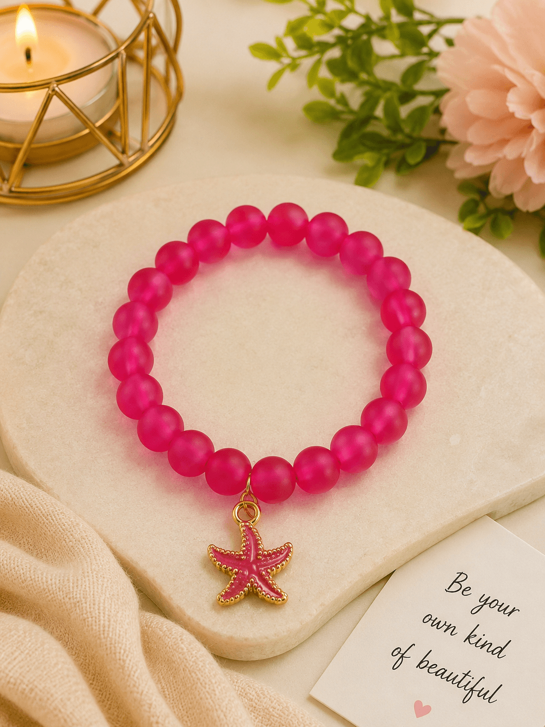 Handmade hot pink jelly bead stretch bracelet with a starfish charm resting on a white marble slab next to a candle