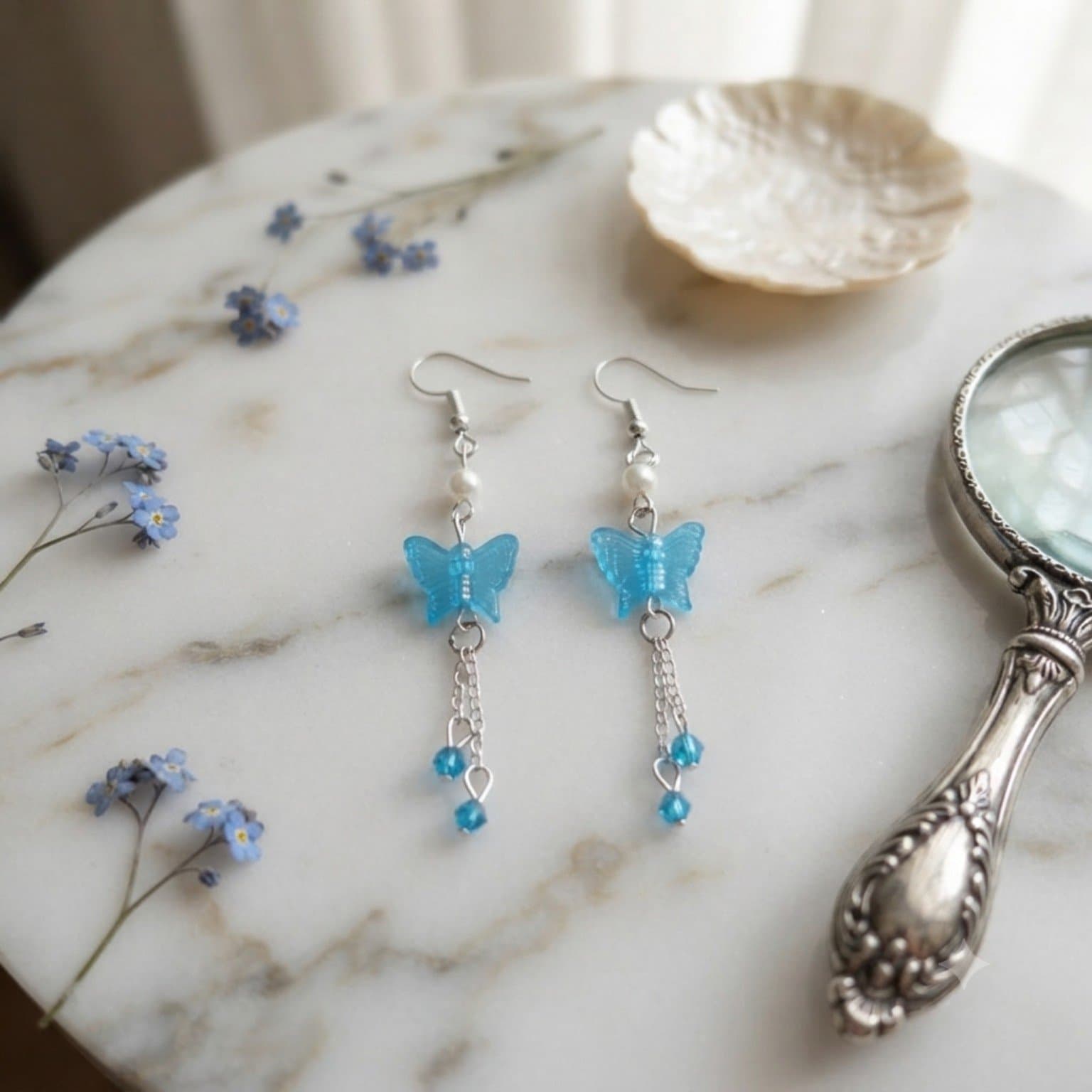 Handmade sky-blue resin butterfly drop earrings with chains and pearls resting on a marble surface next to blue flowers